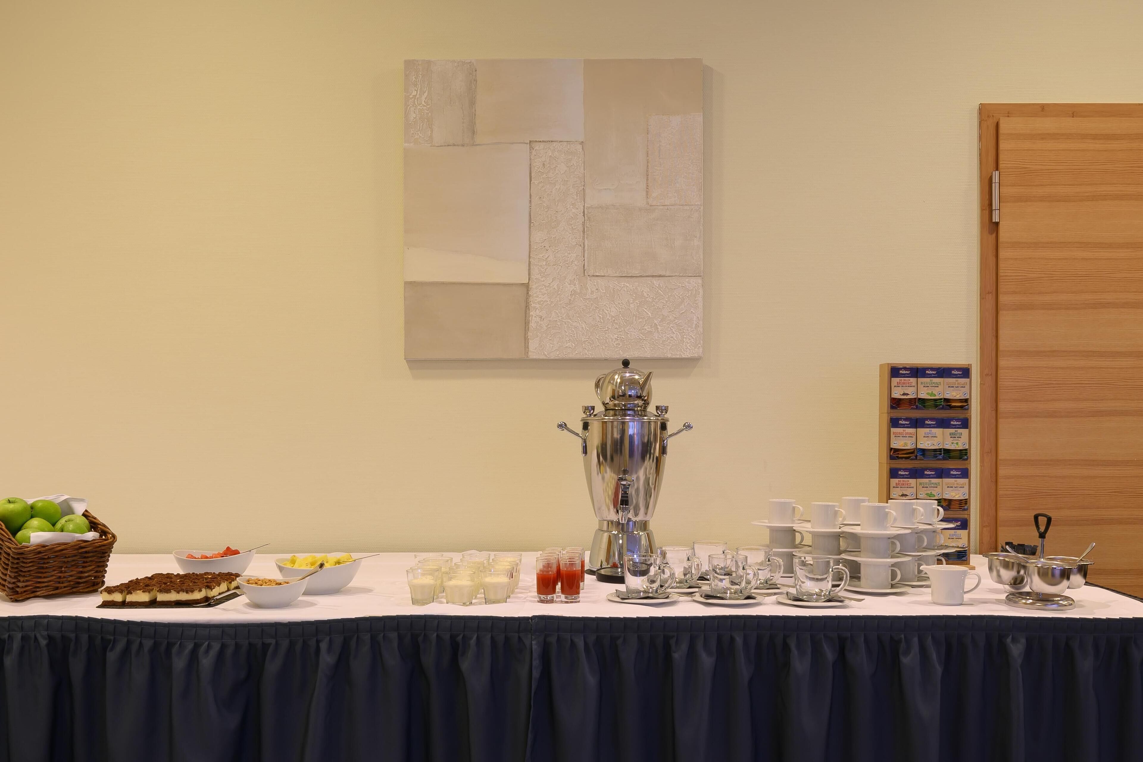 A buffet table with various food items and a large silver urn, likely for hot beverages, against a plain wall with a wooden door and a framed abstract artwork.