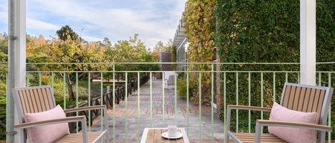 Ein Balkon mit zwei Holzstühlen und einem Tisch, einem Metallgeländer sowie Blick auf einen Garten und ein Gebäude im Hintergrund.