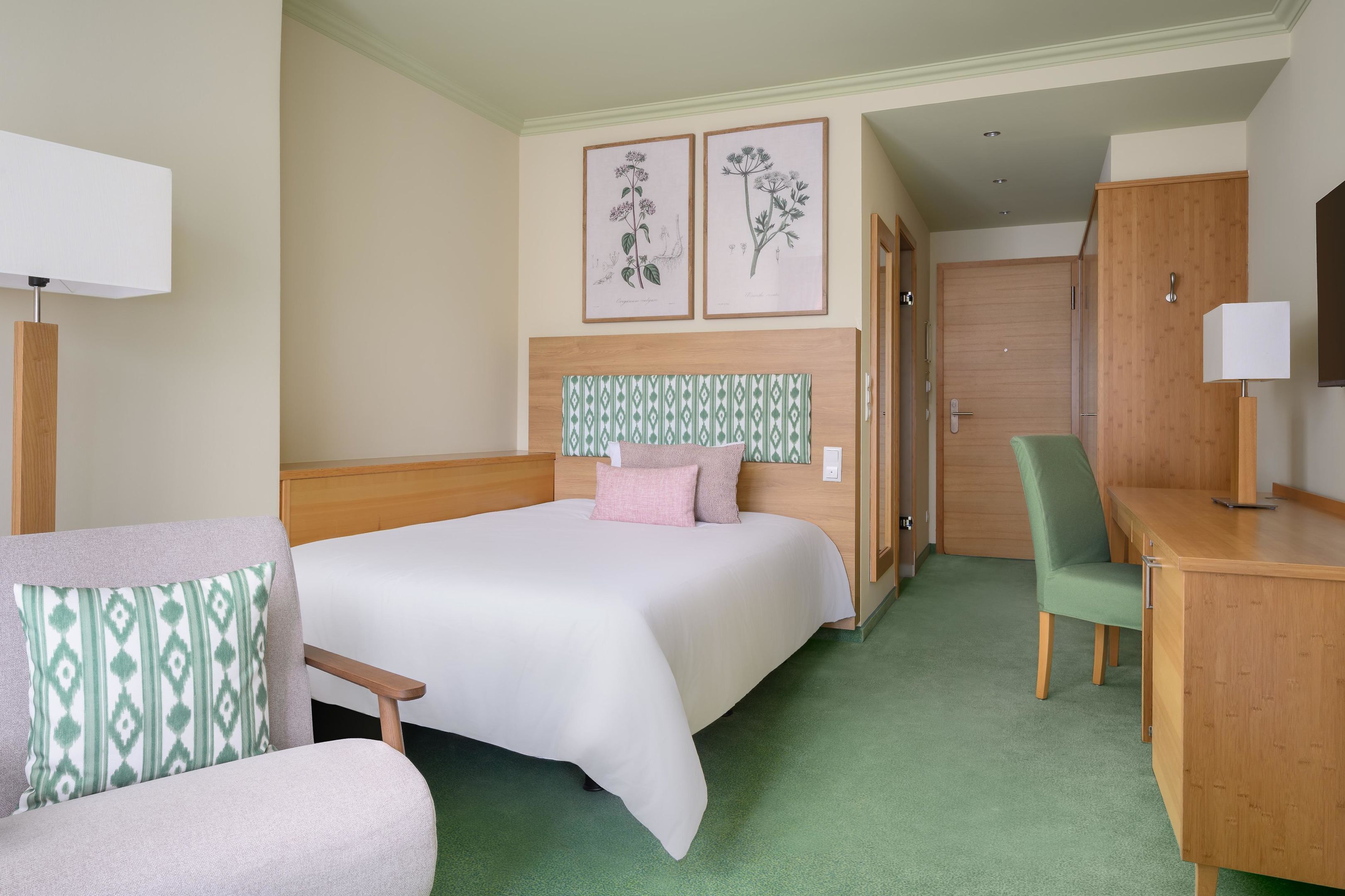 A hotel room with a bed, a desk, a chair, and two framed botanical illustrations on the wall.