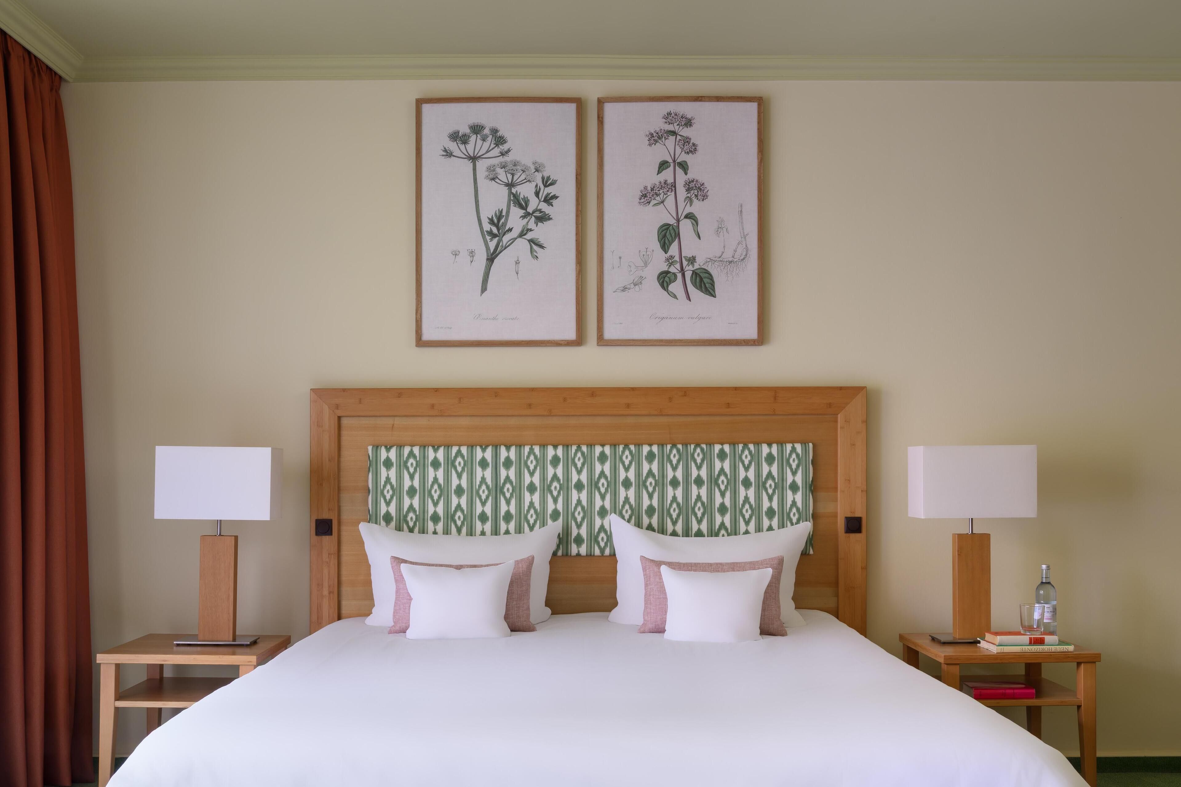 A neatly made double bed with white and pink pillows, a wooden headboard with a patterned wallpaper, and two bedside tables with lamps and books.