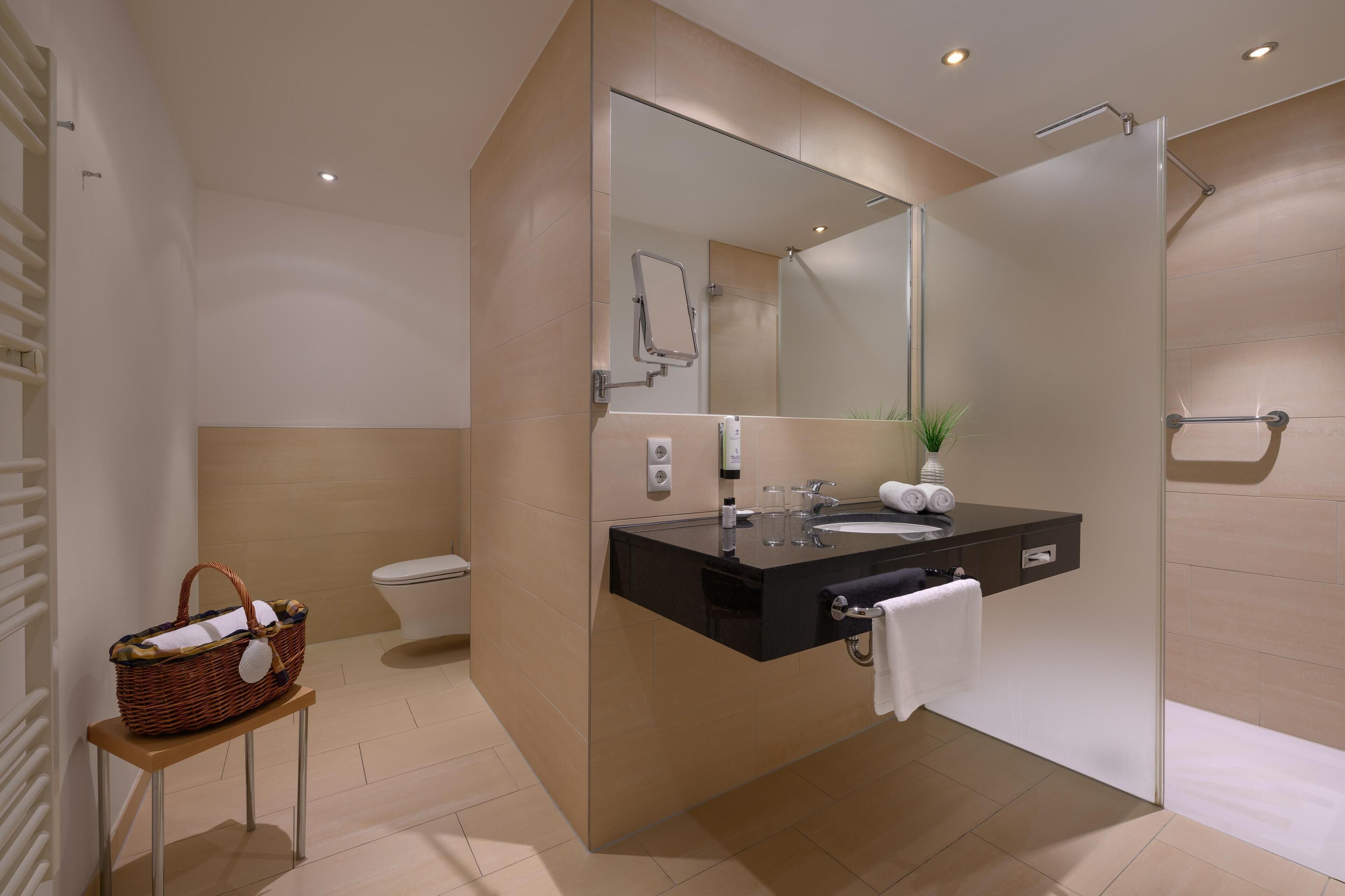 Un baño moderno con cabina de ducha de vidrio, tocador negro con espejo y una cesta de mimbre sobre una mesita.