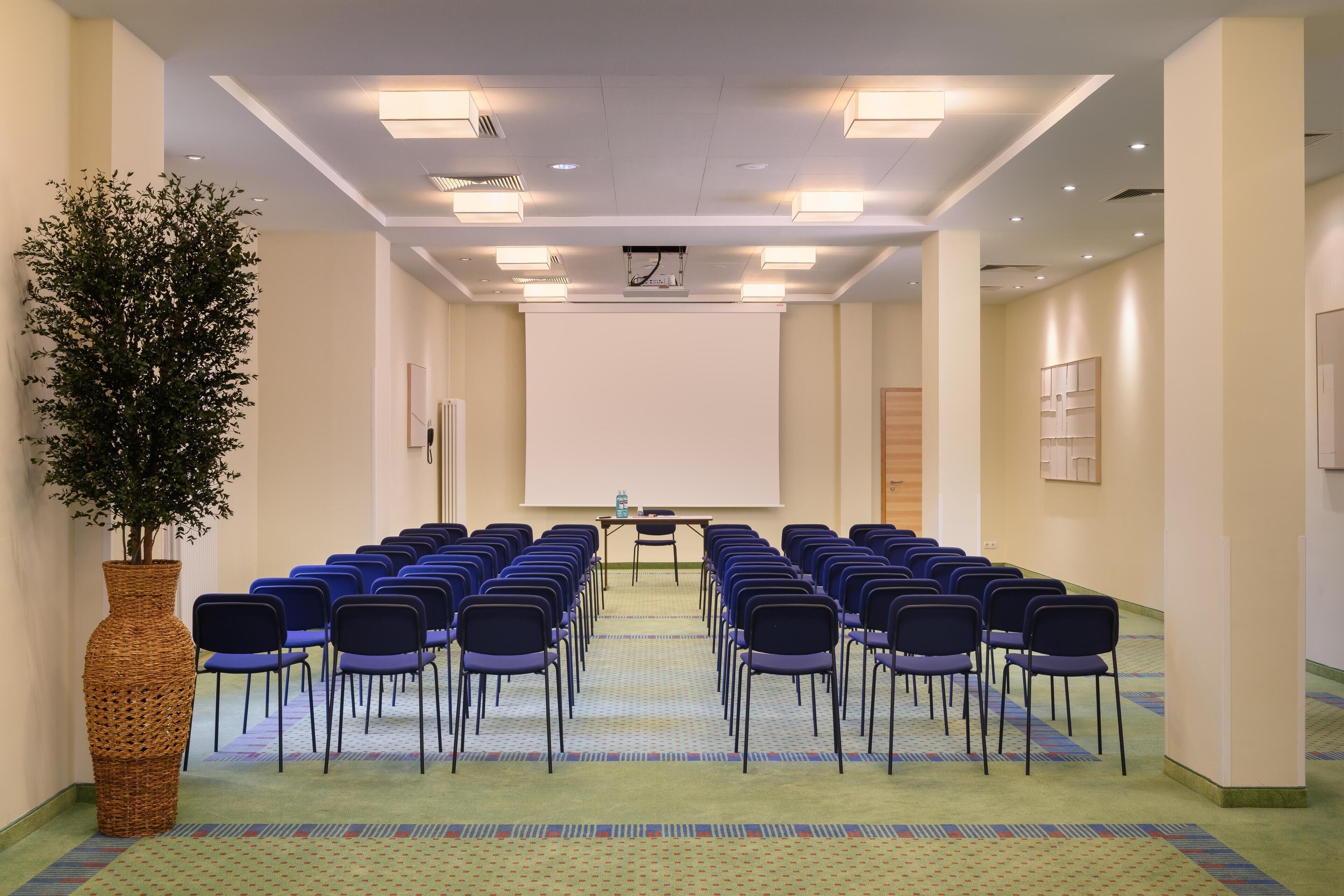 Un salón de conferencias con filas de sillas orientadas hacia una pantalla de proyector, una planta grande y suelo alfombrado.
