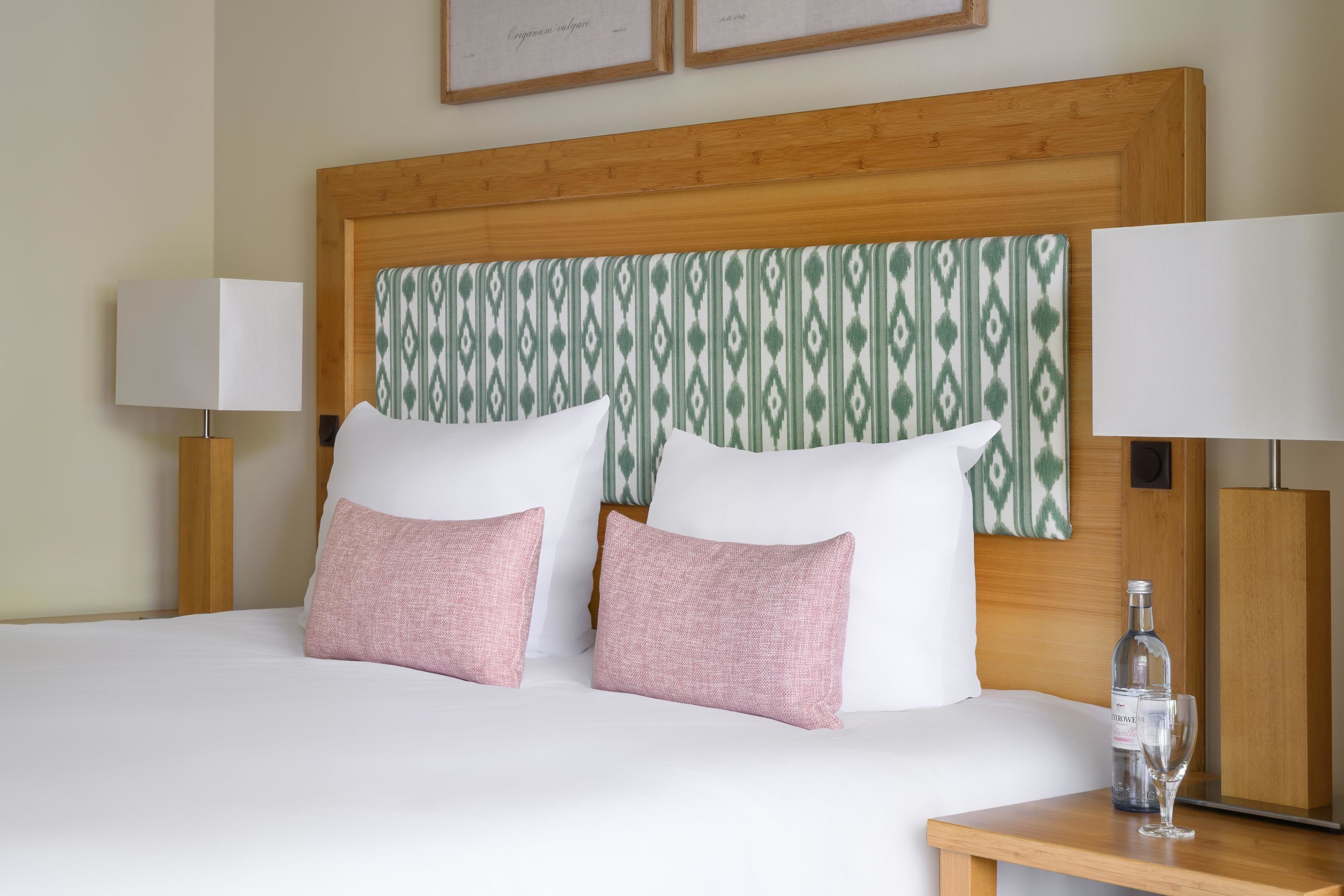 A neatly made bed with white and pink pillows, a wooden headboard, and a bedside table with a bottle and glass.