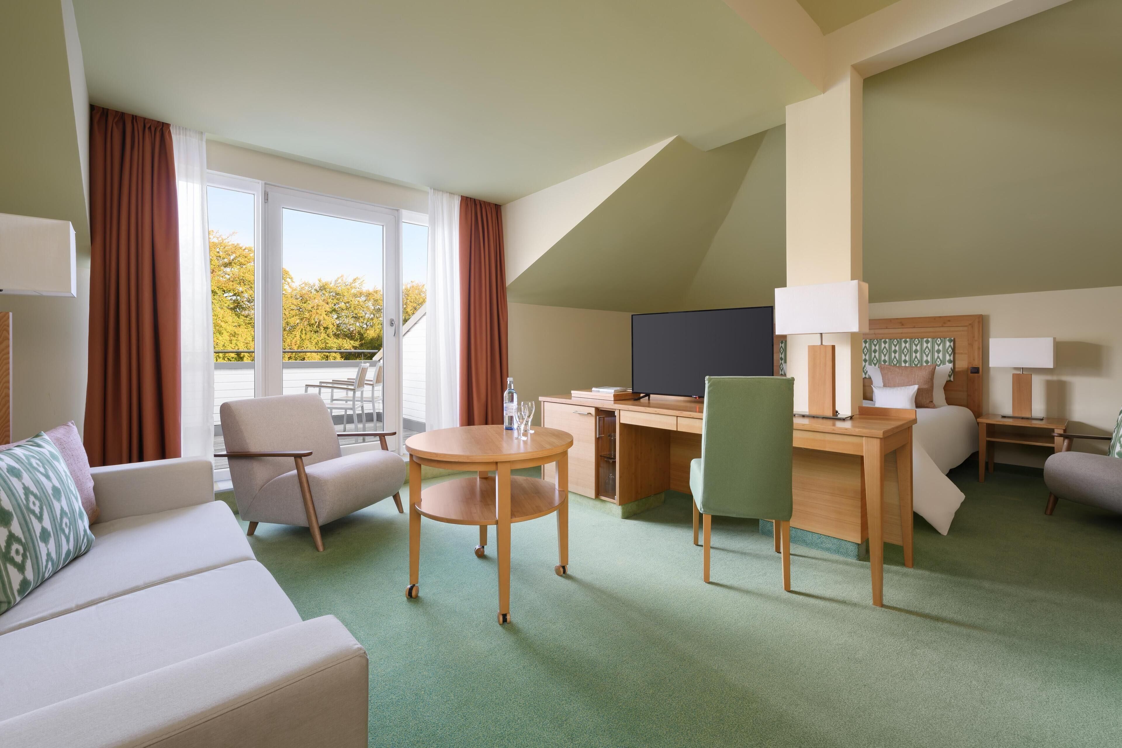 A hotel room with a sofa, two armchairs, a desk, a television, and a balcony with curtains.