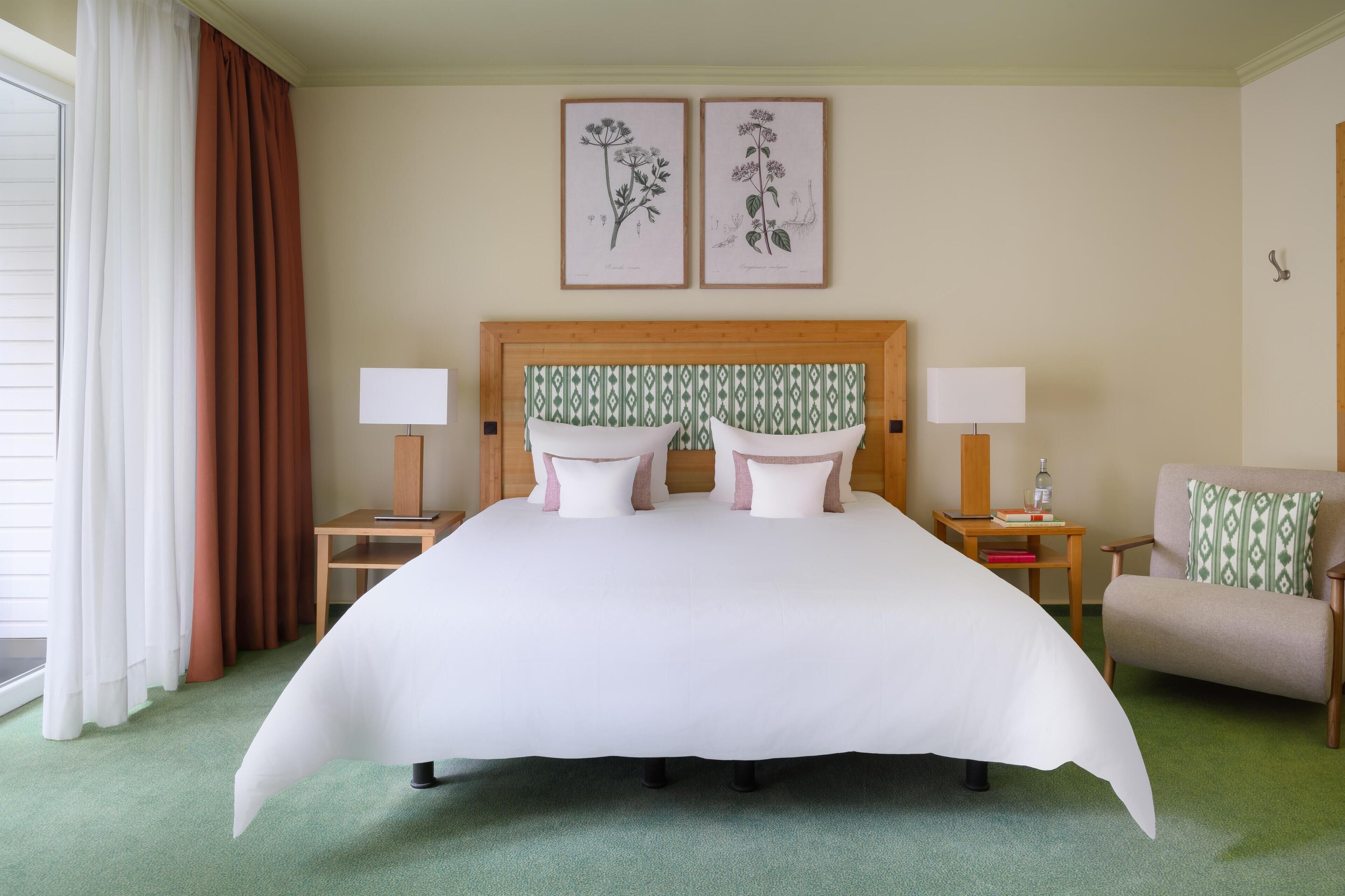 A hotel room with a large bed, two bedside tables with lamps, a chair, and two framed botanical prints on the wall.