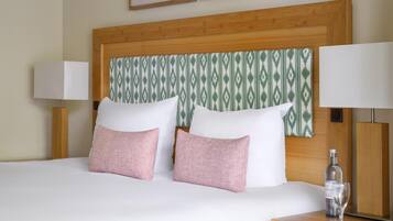 A neatly made bed with two white pillows and two pink pillows, a wooden headboard with a green patterned panel, and a bedside table with a bottle and glass.