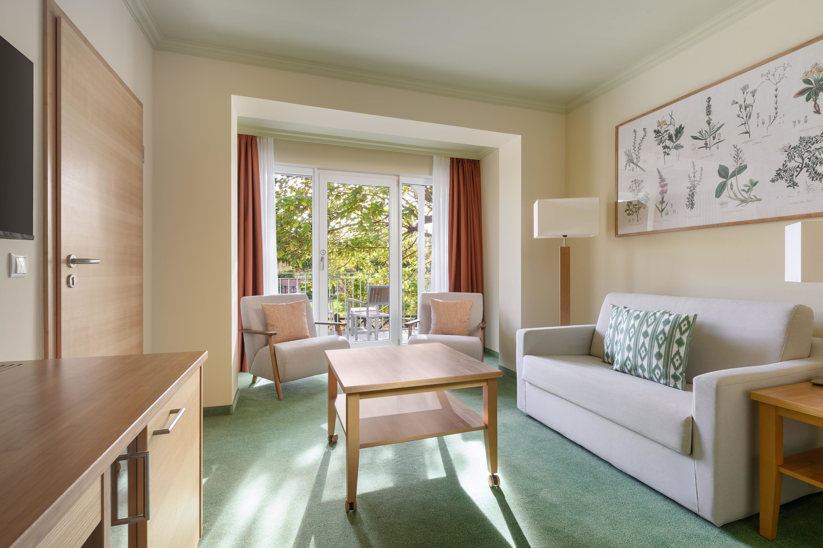 A hotel room with a wooden desk, a white sofa, a green carpet, and a large window with curtains.