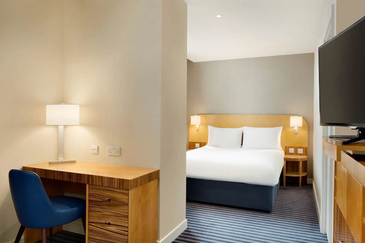 Une chambre d’hôtel avec un bureau en bois, une chaise bleue, un lit avec du linge de maison blanc, deux lampes de chevet et un téléviseur à écran plat.