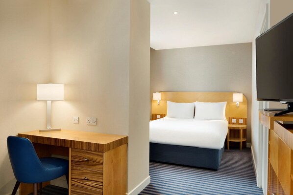 A hotel room with a wooden desk, a blue chair, a bed with white linens, two bedside lamps, and a flat-screen TV.
