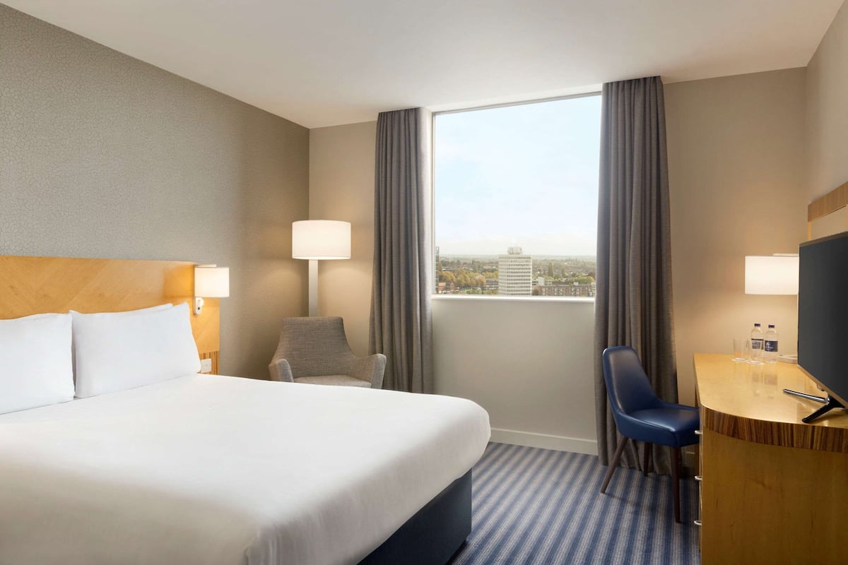 Une chambre d’hôtel avec un grand lit, un bureau, une chaise, une fenêtre avec des rideaux et une vue sur le paysage urbain.