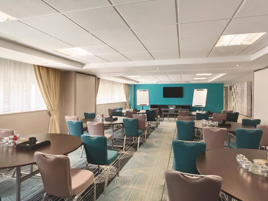 Une salle de conférence aux murs de couleur turquoise, dotée de plusieurs tables et chaises, d’un grand écran et offrant une vue sur la ville grâce à de grandes fenêtres.