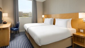 Quarto de hotel com duas camas, uma escrivaninha, uma cadeira e uma janela grande com vista para edifícios.