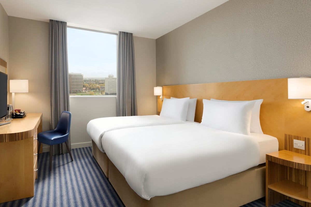 Une chambre d’hôtel avec deux lits, un bureau, une chaise et une grande fenêtre donnant sur des immeubles.