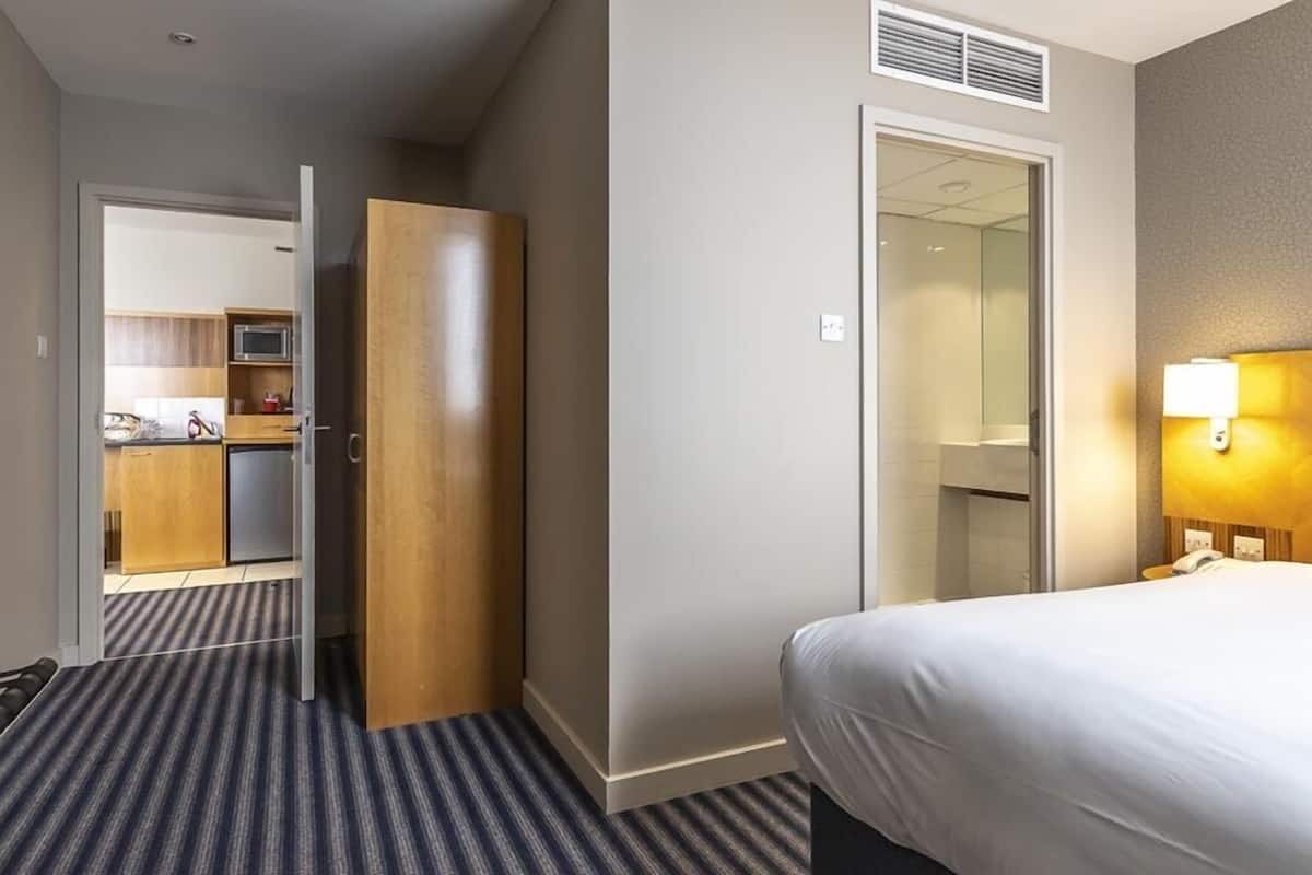 A hotel room with a bed, a wardrobe, a bathroom with a mirror, and a kitchenette in the background.