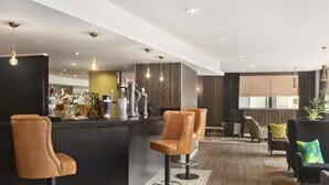Uma área de bar moderna com banquetas de couro, balcão com diversos tipos de garrafas e um espaço de estar com cadeiras e um sofá.