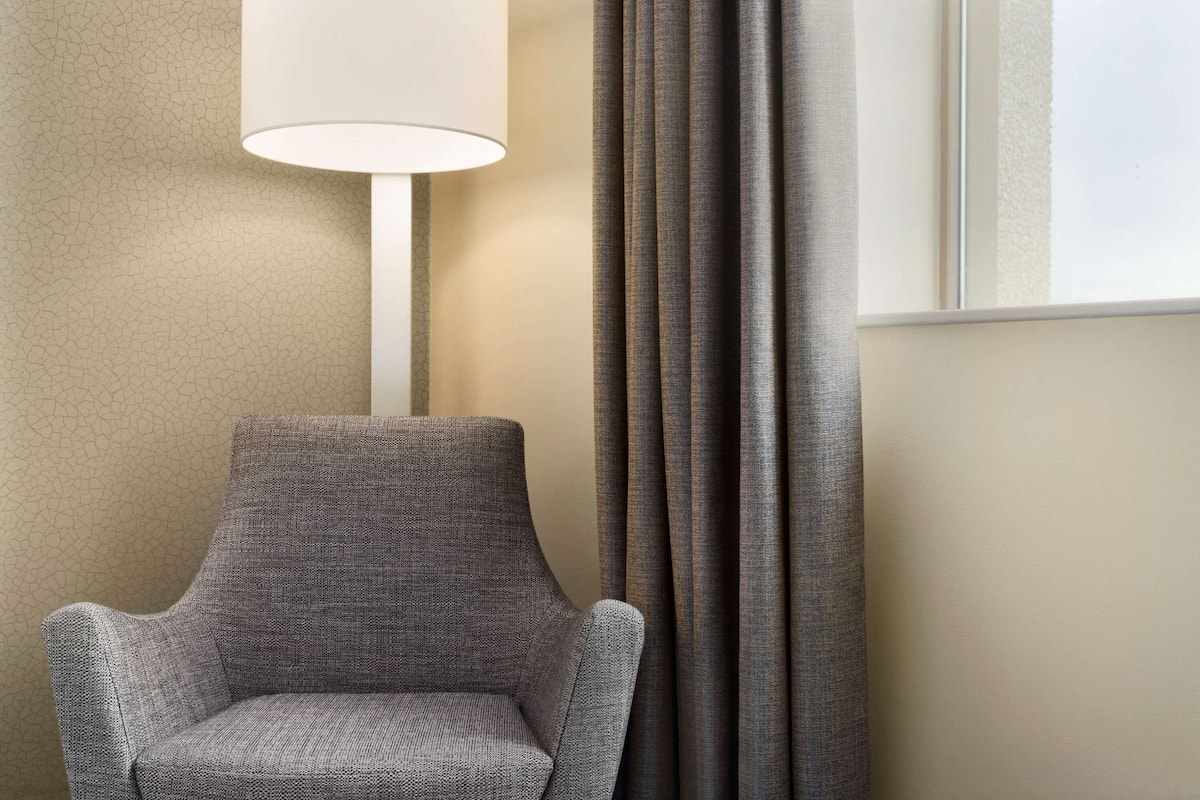 Une chaise recouverte d’un tissu texturé, une lampe avec un abat-jour blanc et une fenêtre avec des rideaux.