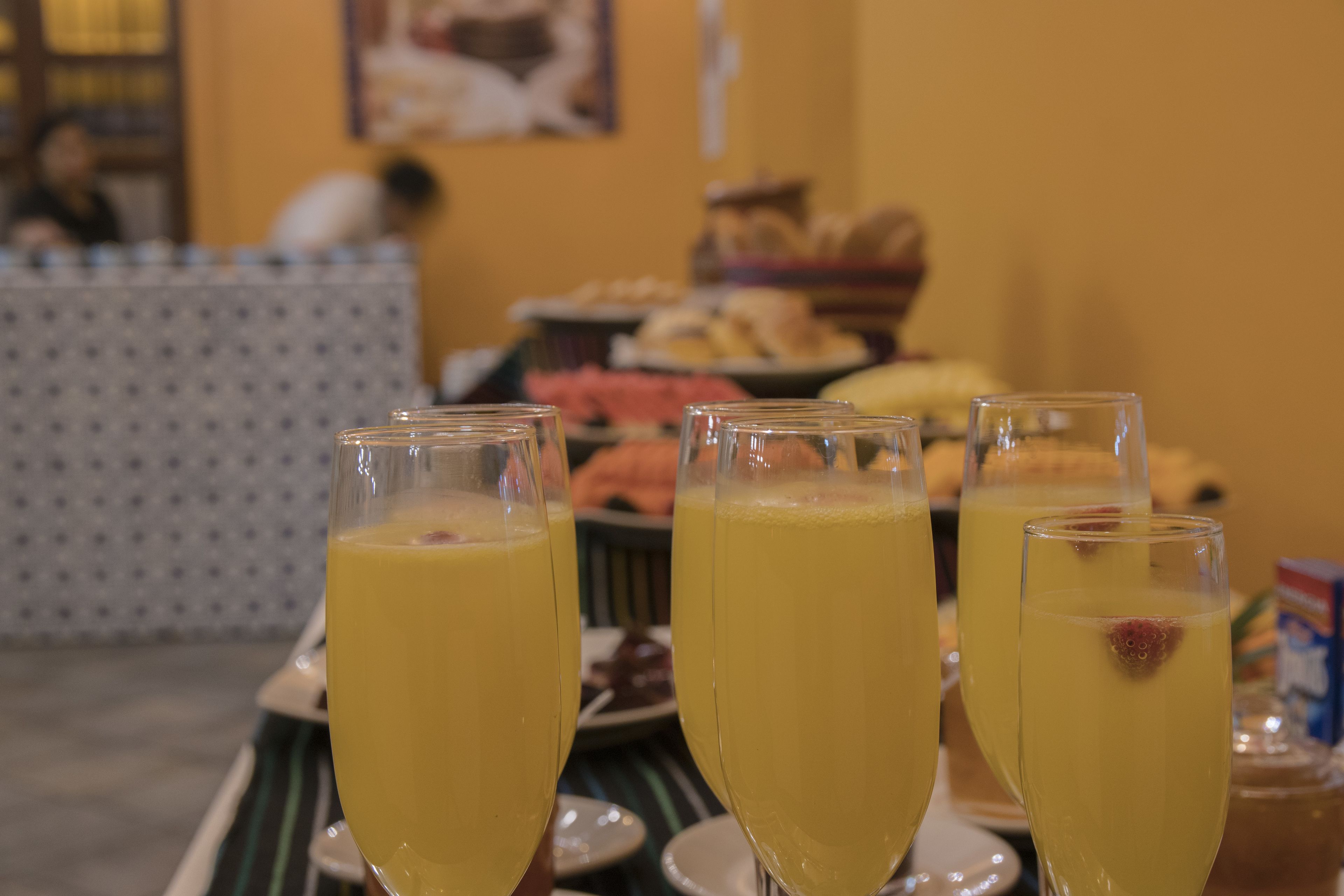 Una mesa con tres vasos de una bebida amarilla, posiblemente un cóctel o jugo, decorada con una fresa. Al fondo, se observa un bufé con diferentes alimentos y una pared con diseño.