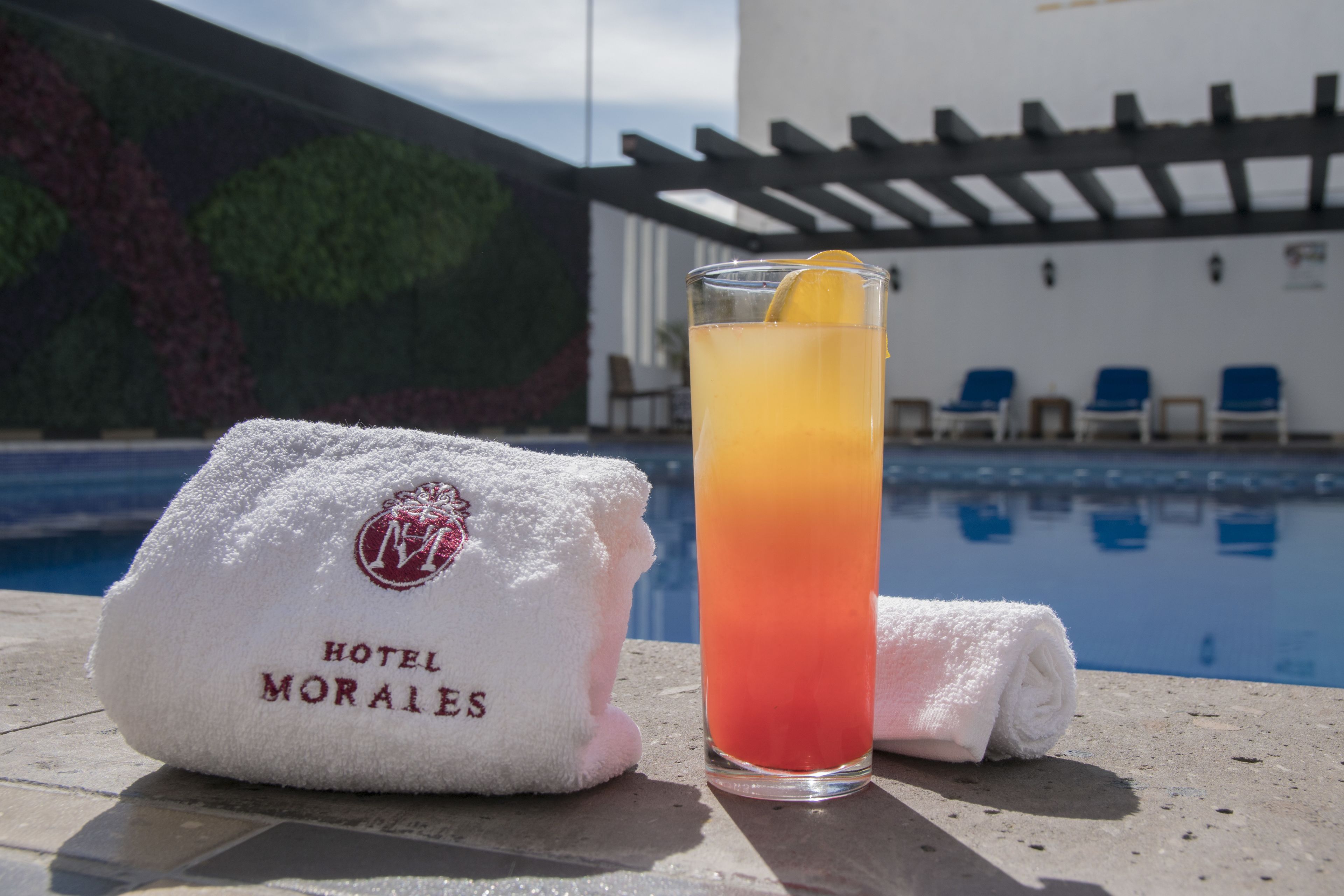 Un vaso de cóctel con una rodaja de limón sobre una mesa junto a la piscina, una toalla blanca con el logotipo de 'HOTEL MORALES' y una piscina con sillas azules al fondo.