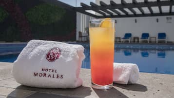 A glass of cocktail with a lemon slice on a poolside table, a white towel with "HOTEL MORALES" logo, and a pool with blue chairs in the background.
