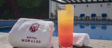 Un vaso de cóctel con una rodaja de limón sobre una mesa junto a la piscina, una toalla blanca con el logotipo de 'HOTEL MORALES' y una piscina con sillas azules al fondo.