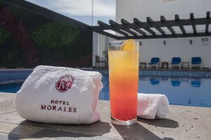 A glass of cocktail with a lemon slice on a poolside table, a white towel with "HOTEL MORALES" logo, and a pool with blue chairs in the background.