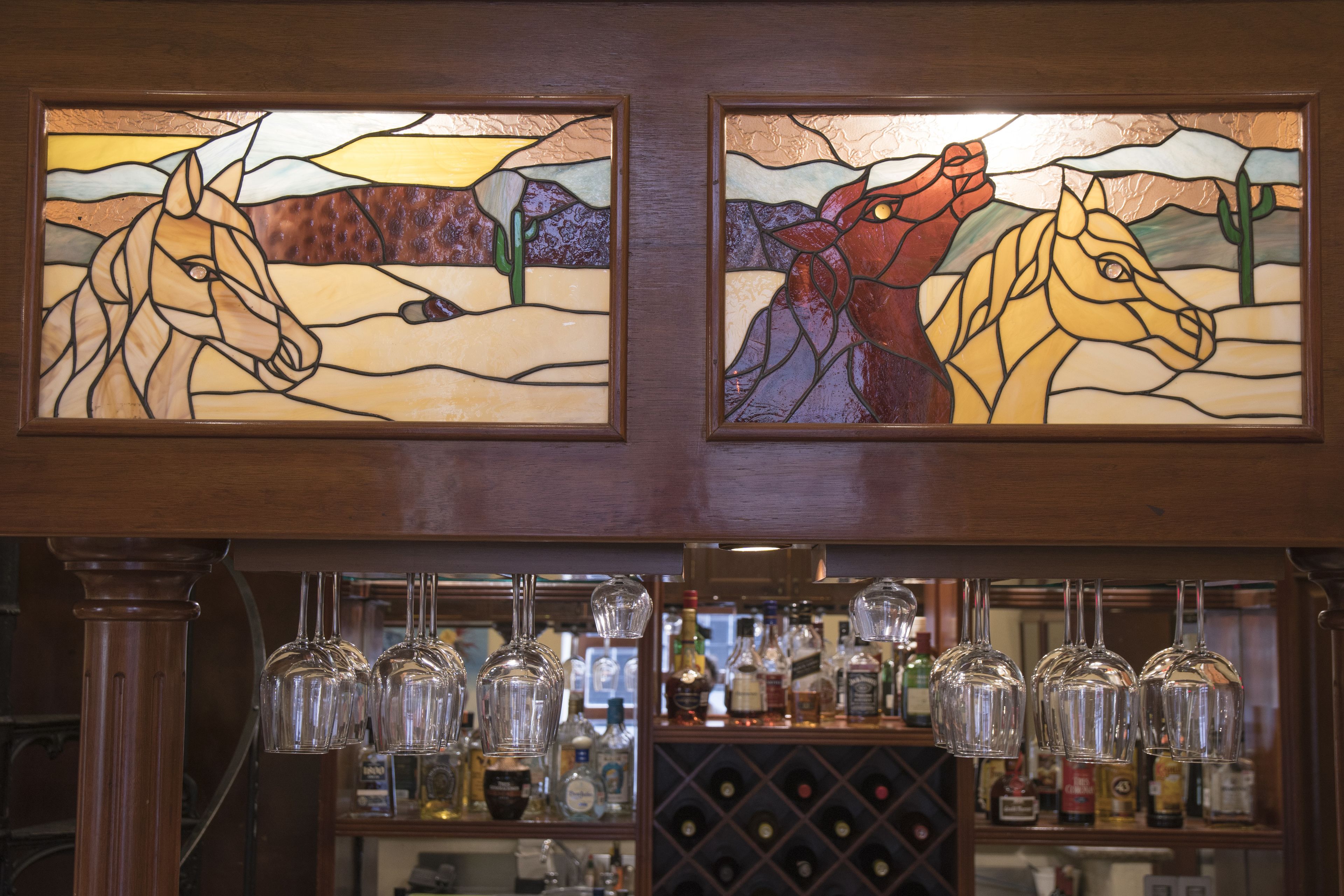 Dos cuadros enmarcados con pinturas de caballos de vidrio esmaltado sobre una zona de bar, con copas de vino y botellas.