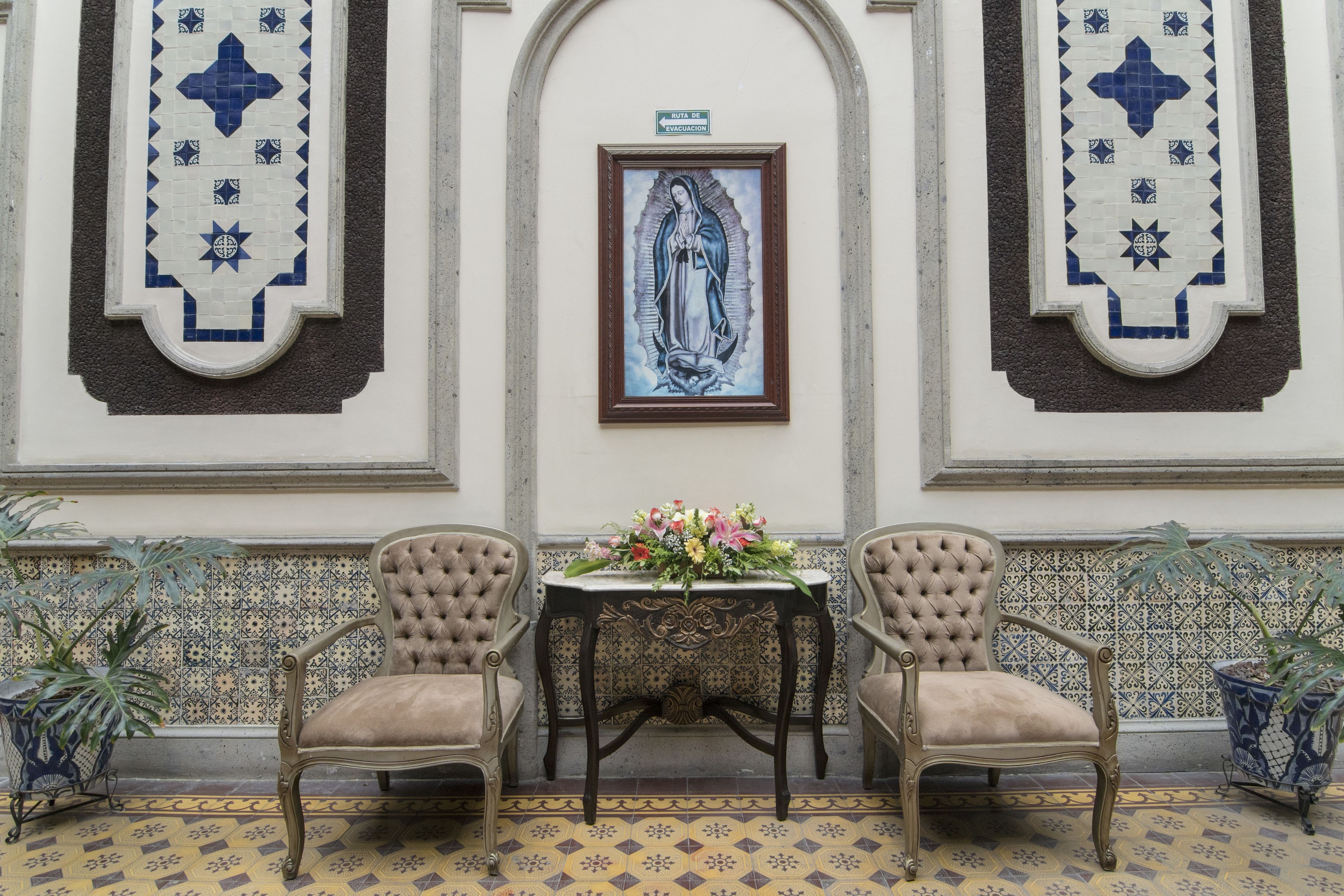 A room with two chairs, a table with a floral arrangement, and a framed religious painting on the wall.