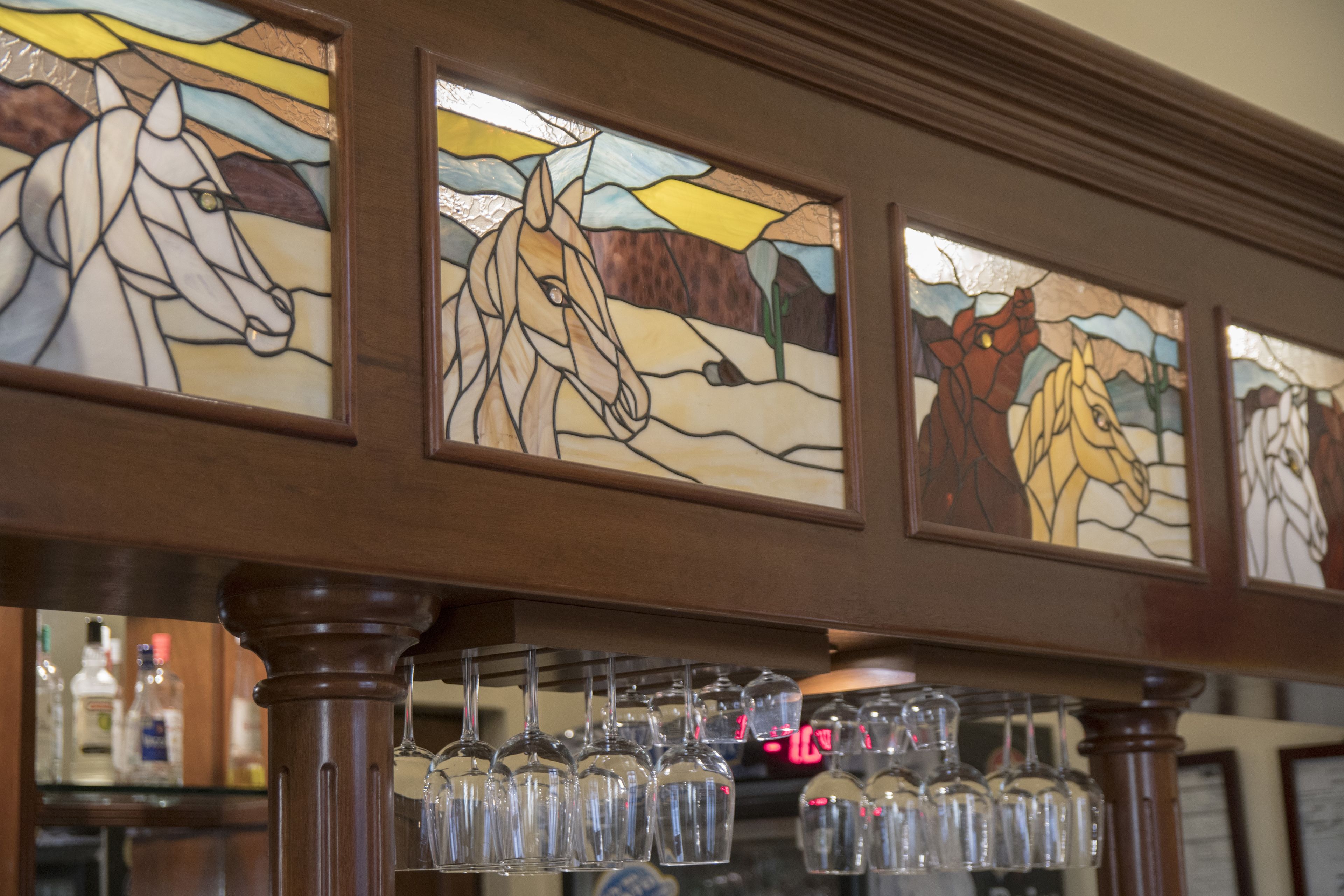 A bar area with stained glass windows depicting horses, a wooden bar counter, and hanging wine glasses.