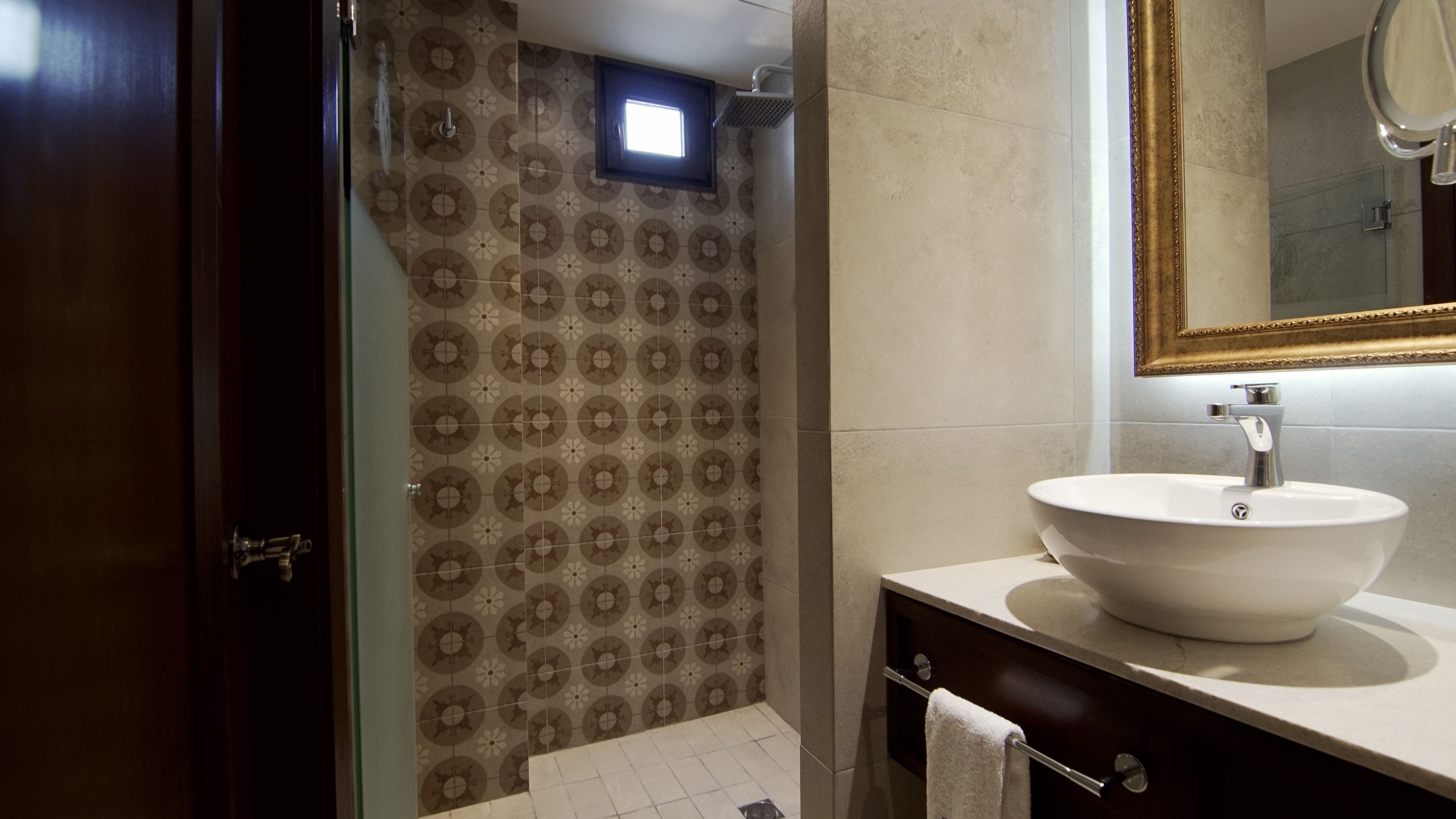 Un baño con pared de ducha con diseño, un lavamanos blanco y un espejo con marco dorado.