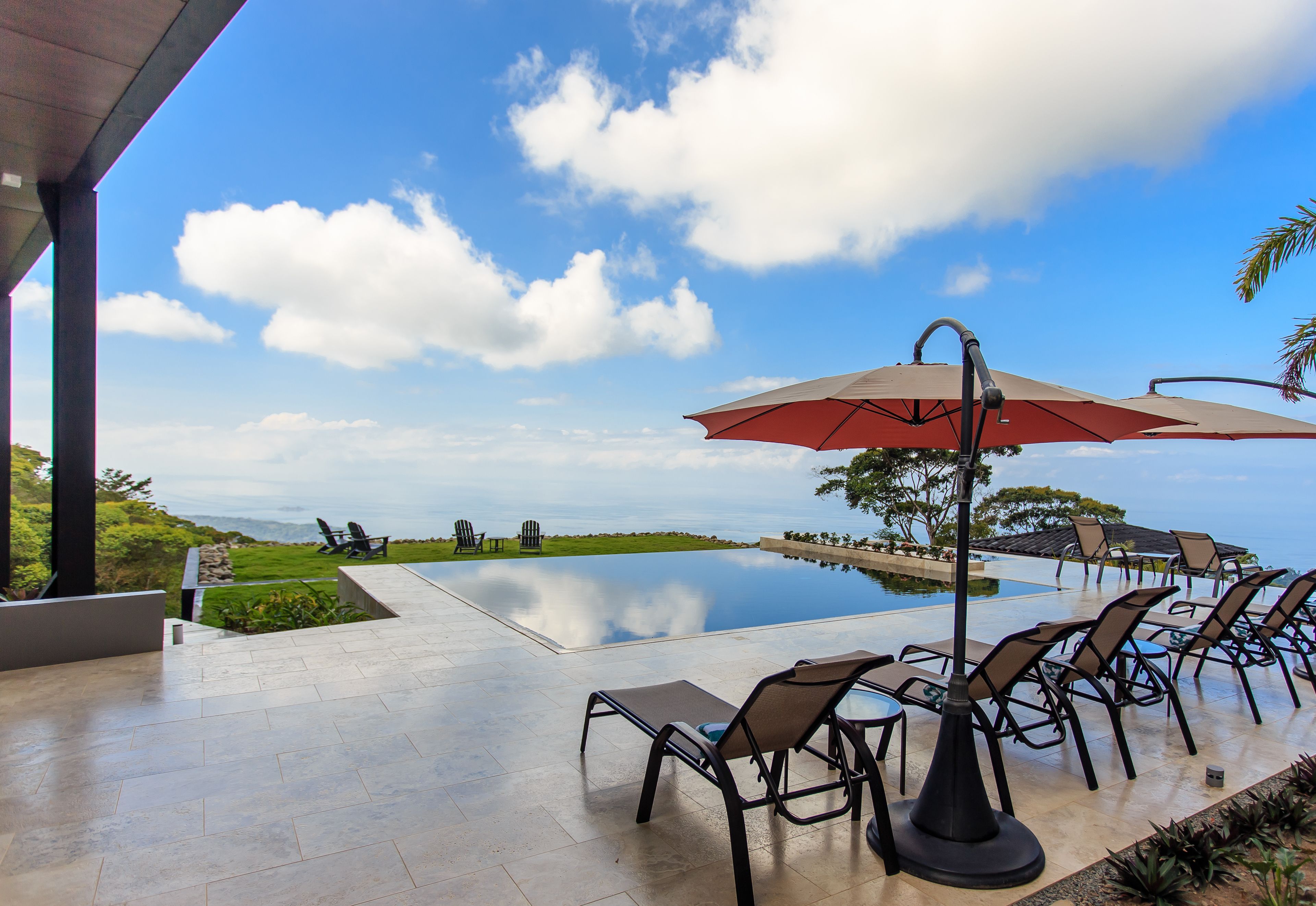 Una piscina infinita con sillones de descanso y sombrillas, cielo despejado y vista al océano.