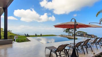 Una piscina infinita con sillones de descanso y sombrillas, cielo despejado y vista al océano.