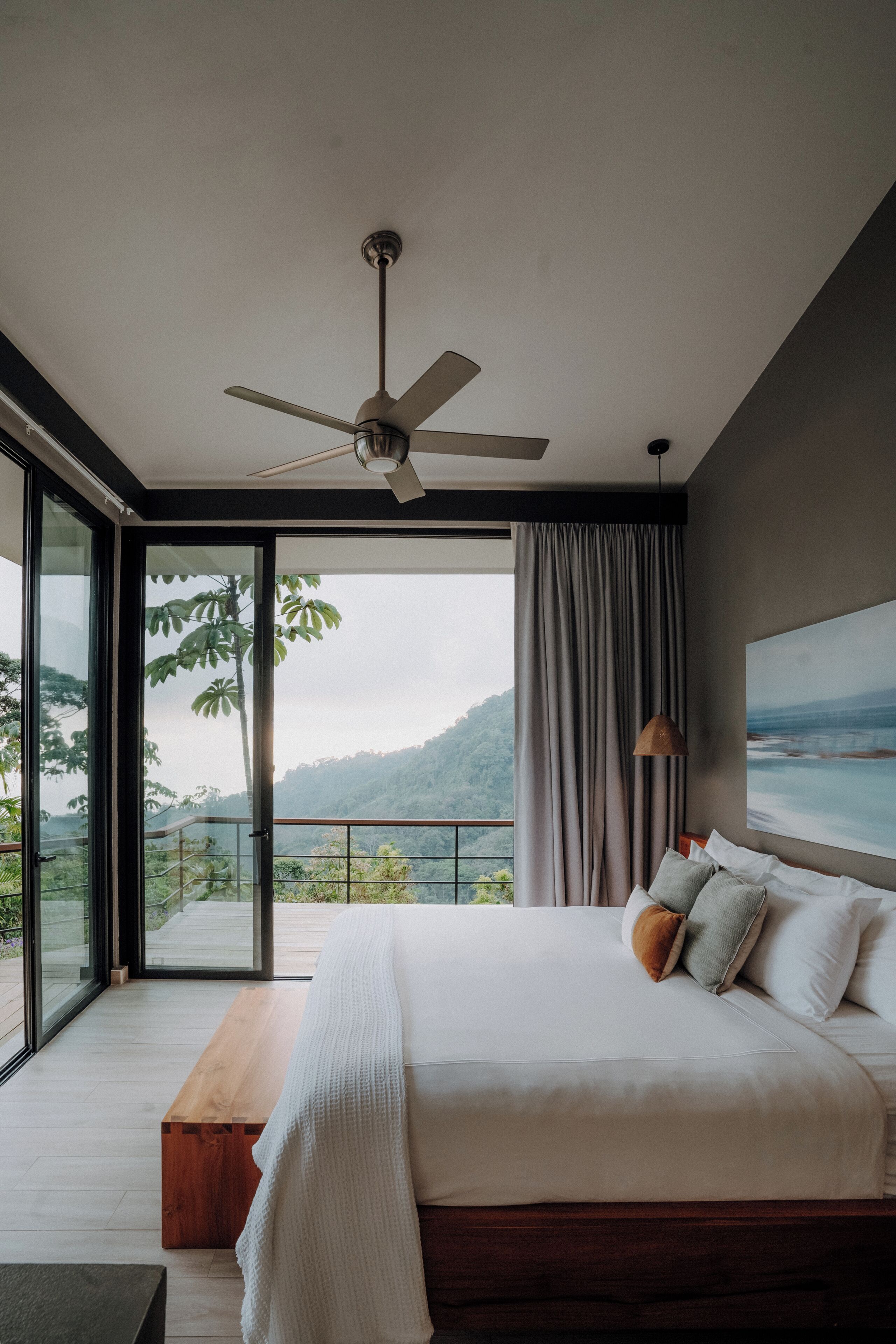 A bedroom with a large bed, a ceiling fan, a view of a mountainous landscape through a sliding glass door, and a wooden bench.