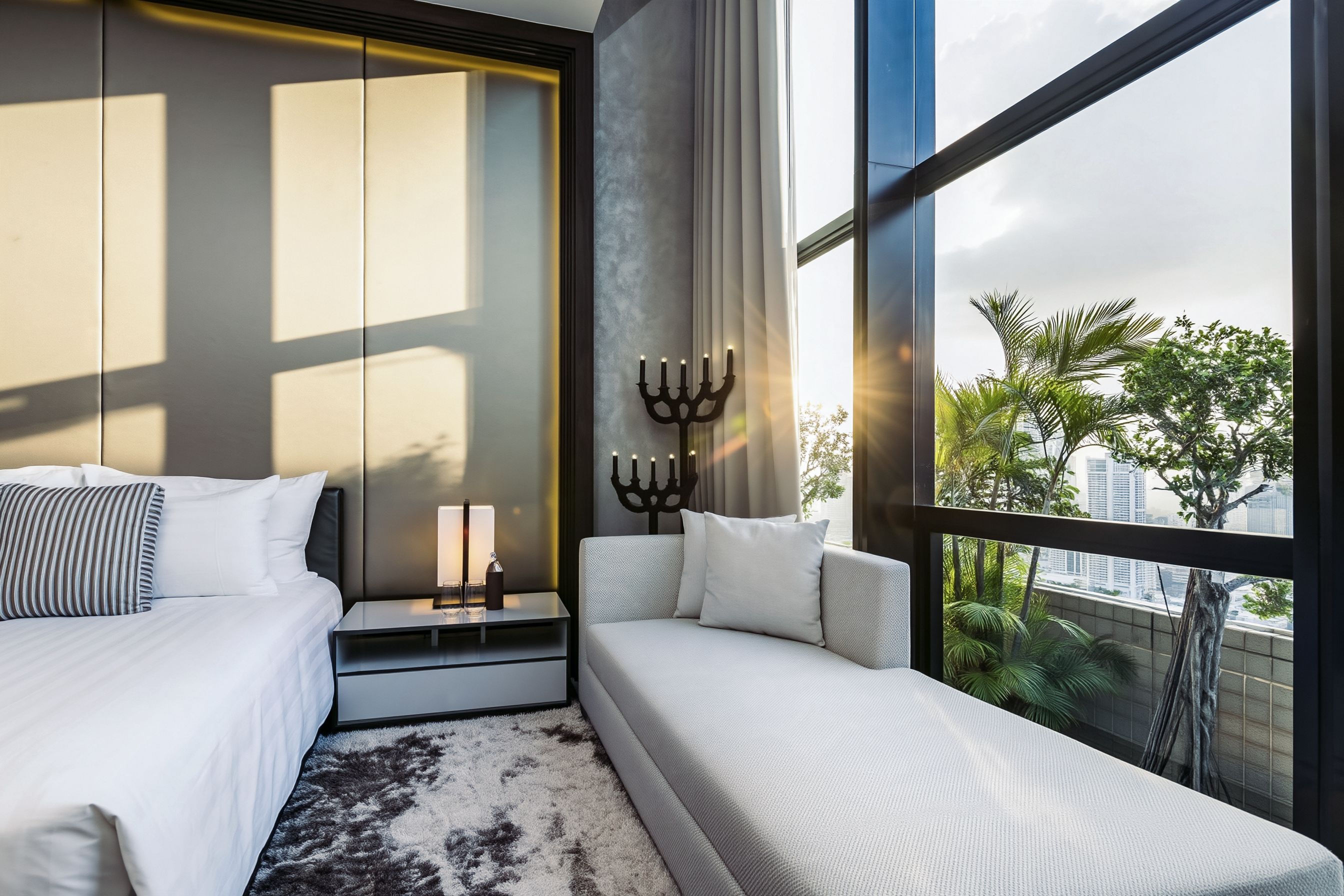 Quarto de hotel moderno com uma janela grande, uma cama, uma mesa de cabeceira com luminária e um objeto decorativo na parede.
