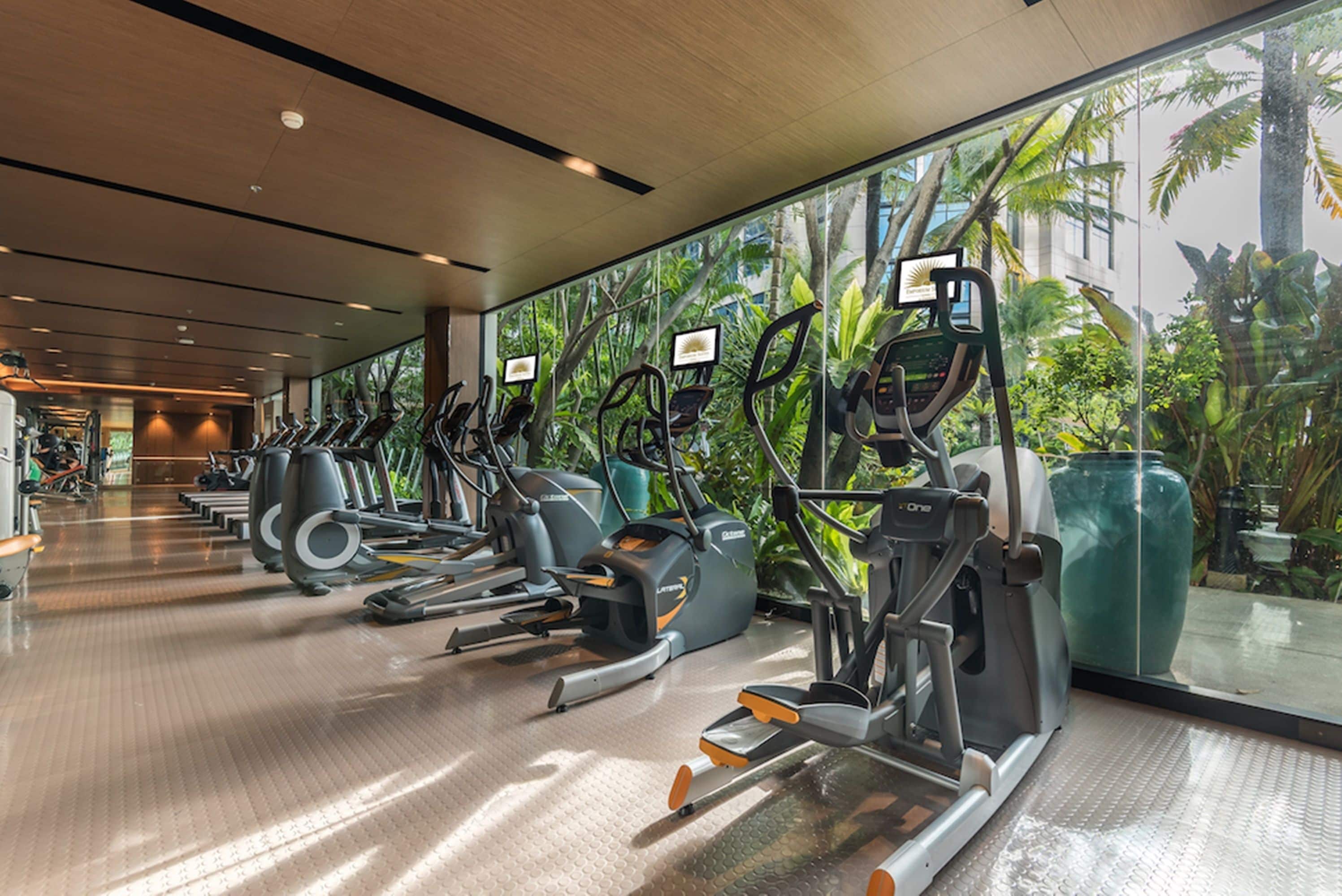 Une salle de sport équipée de tapis de course, de vélos elliptiques et de vélos d’appartement, avec de grandes fenêtres offrant une vue sur un paysage tropical.