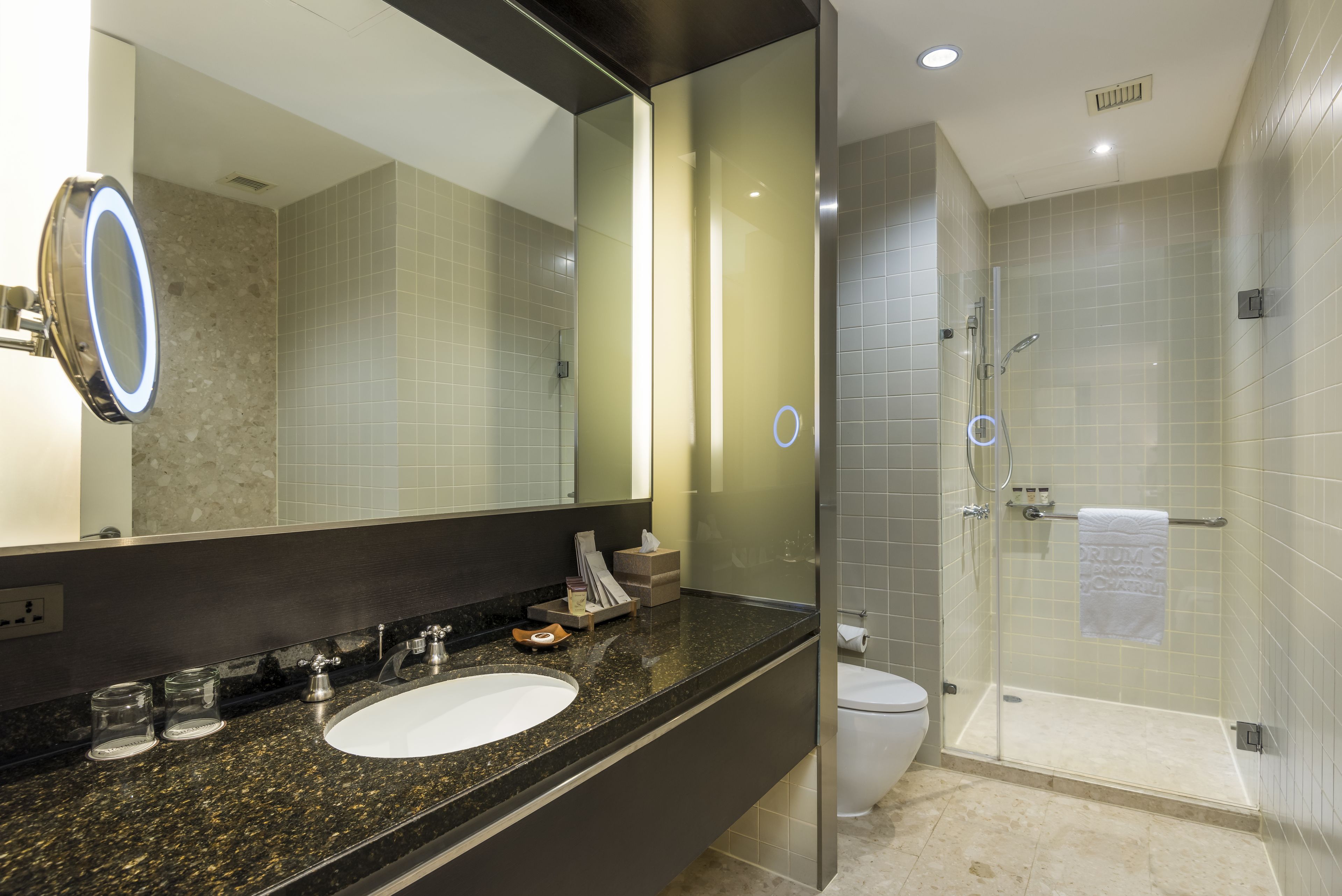 Un baño moderno con encimera de granito, lavamanos blanco, un espejo grande y zona de ducha con paredes de vidrio.