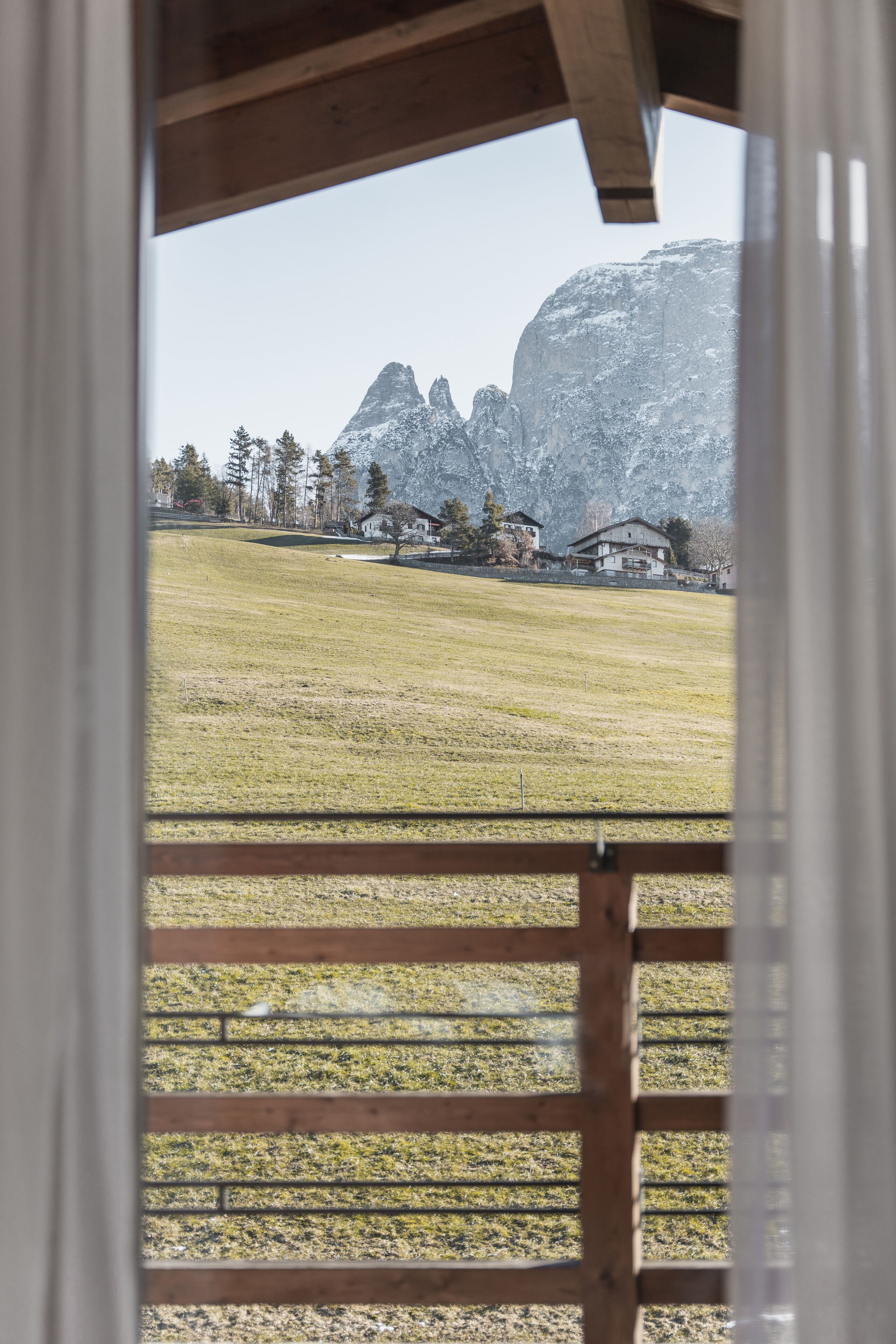 A view through a window with a wooden frame, overlooking a grassy field and a mountain range with a few buildings in the distance.