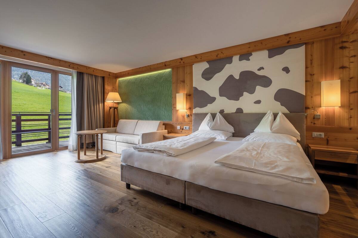 A bedroom with a large bed, a sofa, a small table, and a view of a green field through a sliding door.