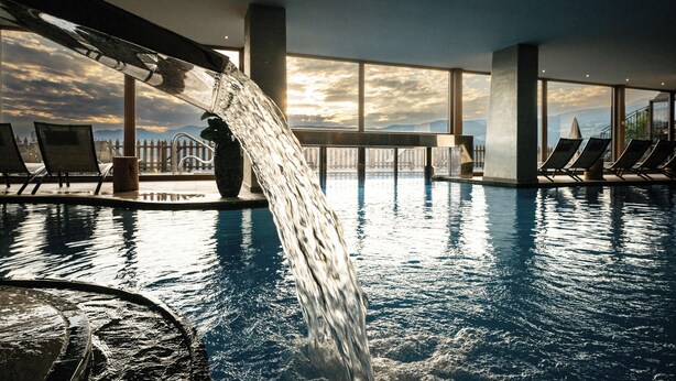 Una piscina con cascada de agua, rodeada de sillones de descanso y grandes ventanales que ofrecen vistas al cielo.
