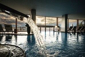 En swimmingpool med vand, der falder ned i den, omgivet af liggestole og store vinduer med udsigt til himlen.