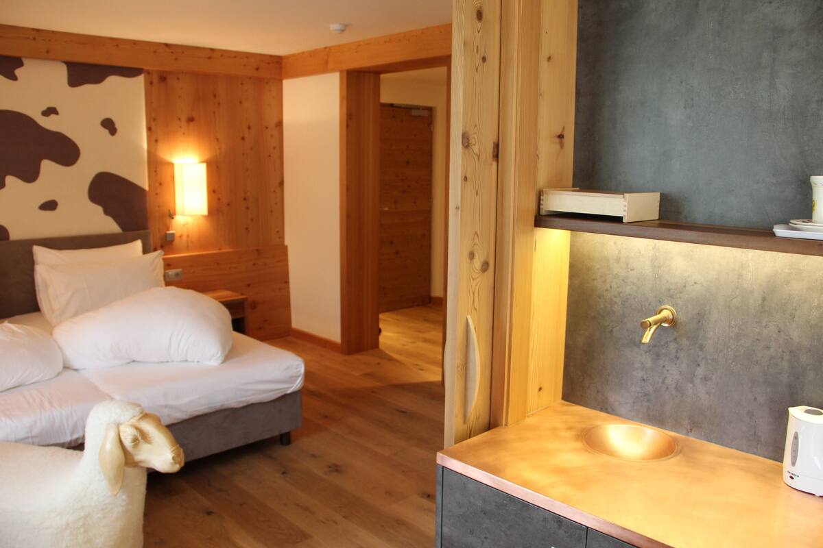 A bedroom with a bed, a wooden headboard, a nightstand, a wall-mounted lamp, and a bathroom with a sink and mirror.
