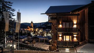 A wooden building with a balcony and a lit interior, surrounded by snow and a town in the background.