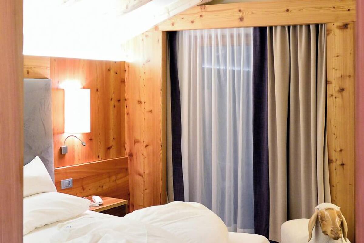 A cozy bedroom with a wooden ceiling, a bed with white linens, a nightstand, a lamp, and a fluffy white sheep decoration.