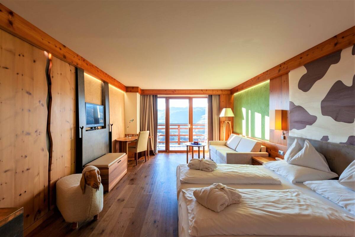 A hotel room with wooden walls, a large bed, a desk, a chair, a sofa, and a view of the mountains through the balcony doors.