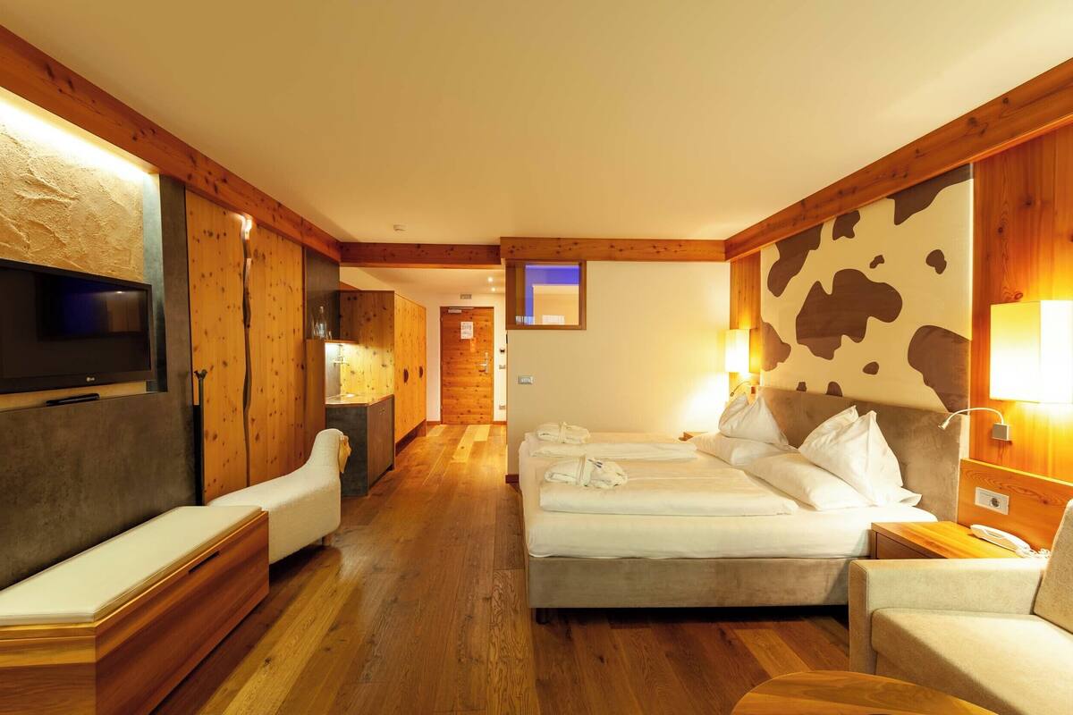 A hotel room with a wooden floor, a bed with white linens, a sofa, a TV mounted on the wall, and a wooden headboard with a cow print design.
