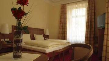 Una habitación de hotel con cama, mesita de noche, lámpara, silla, mesa y un florero con flores.