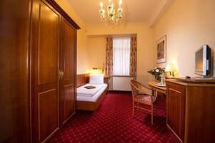 A hotel room with a bed, a desk with a television, a chair, a wardrobe, and a chandelier.