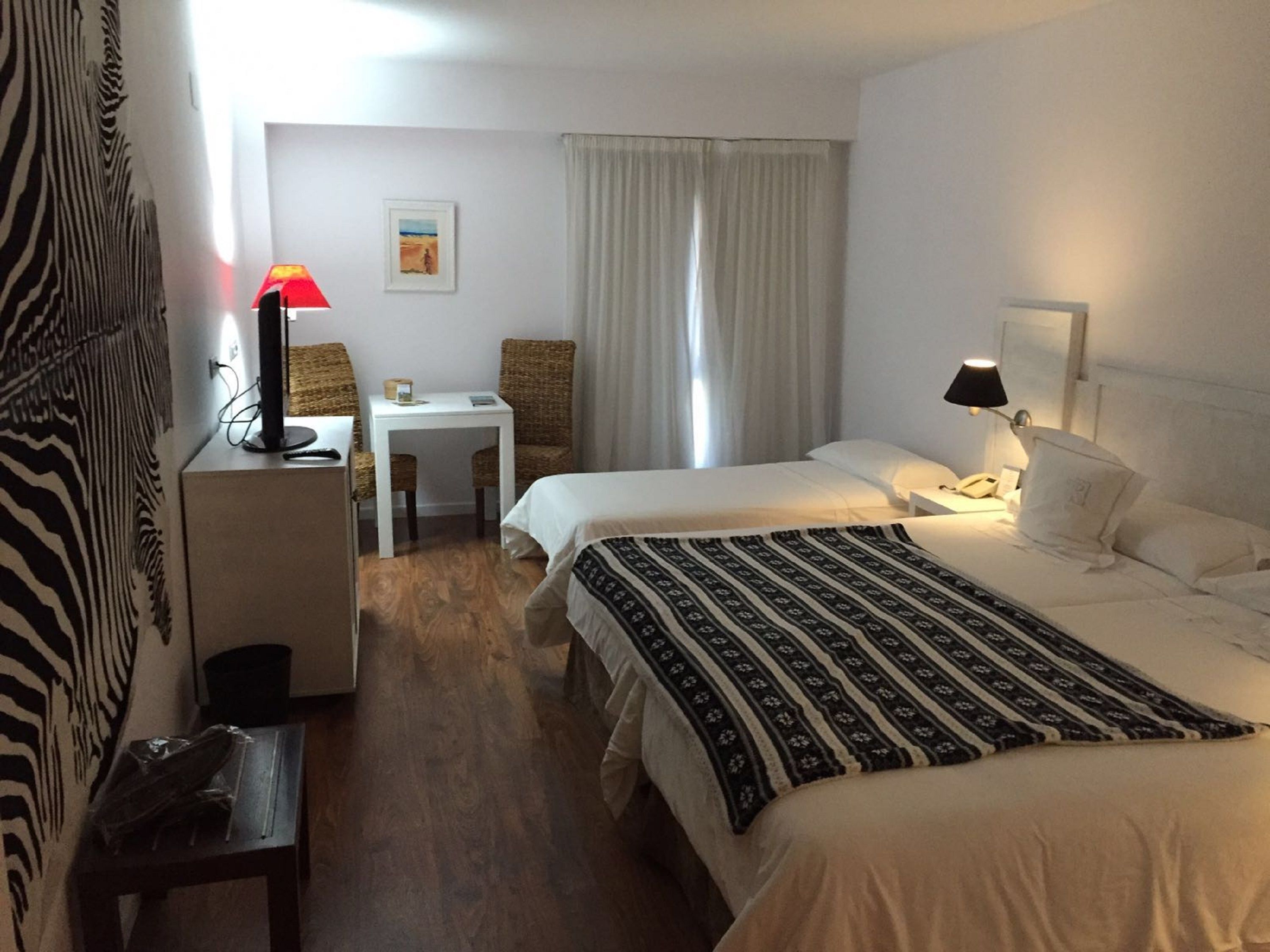 A hotel room with two beds, a small desk, a chair, a lamp, and a painting on the wall.