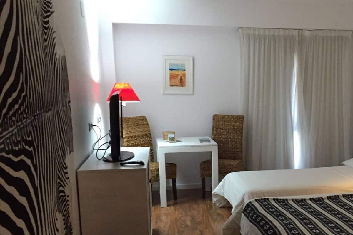 A hotel room with a bed, a desk with a lamp, a chair, a small table, and a painting on the wall.