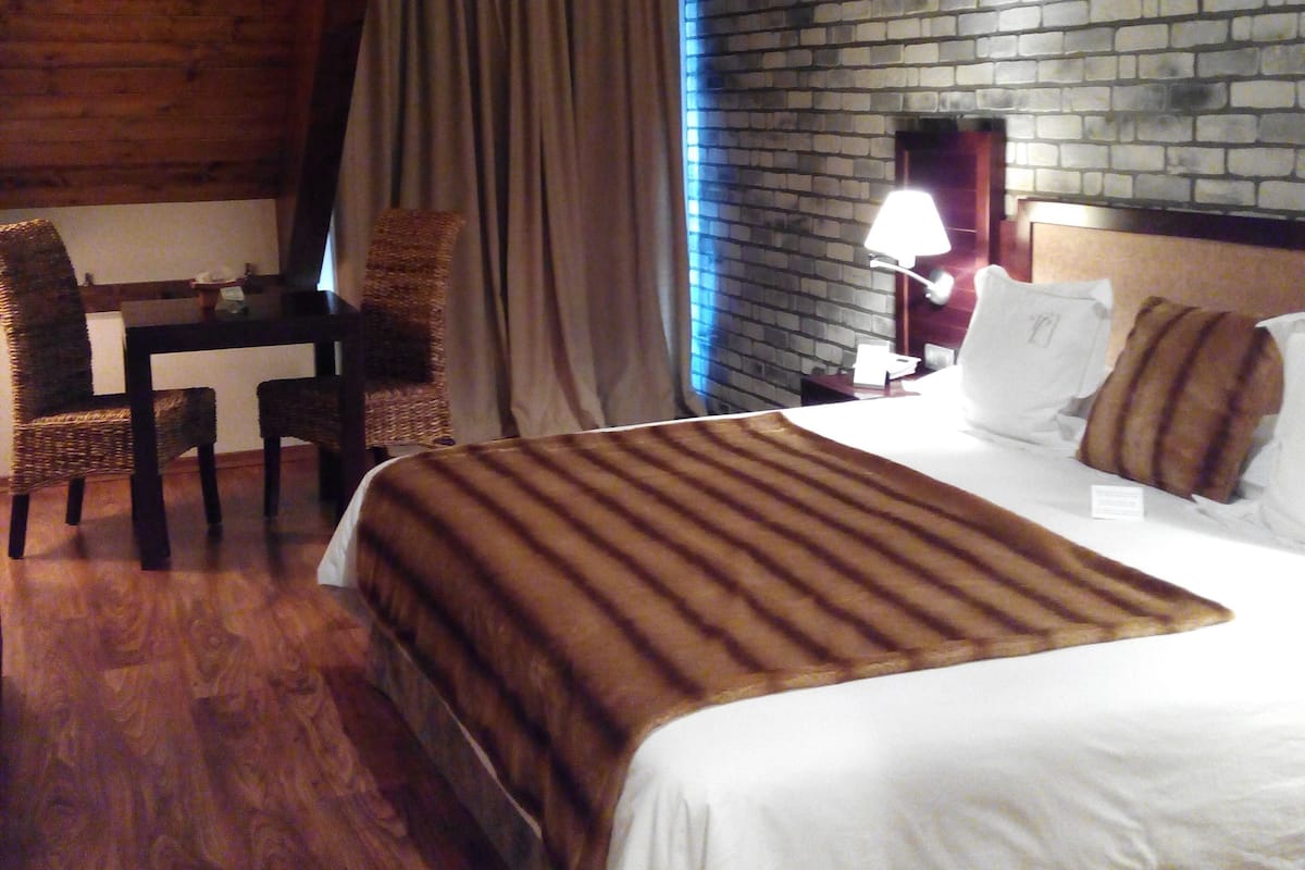 A hotel room with a wooden ceiling, a brick wall, a bed with white bedding, a table with chairs, and a lamp.