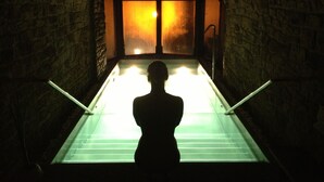Una persona de pie en una habitación tenuemente iluminada, con paredes de piedra y un ventanal grande iluminado.